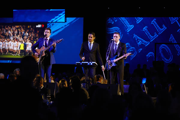 The band in suits on a gala stage with guitar and keys, big screens behind and the dinner crowd in silhouette