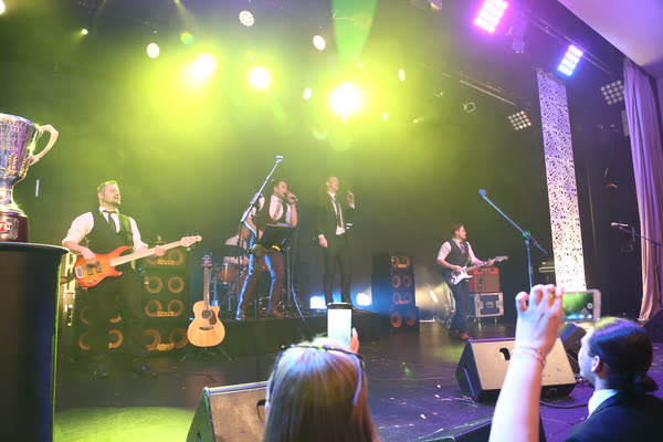 The band on a club stage under green lights, a trophy on the left and fans holding up phones in front