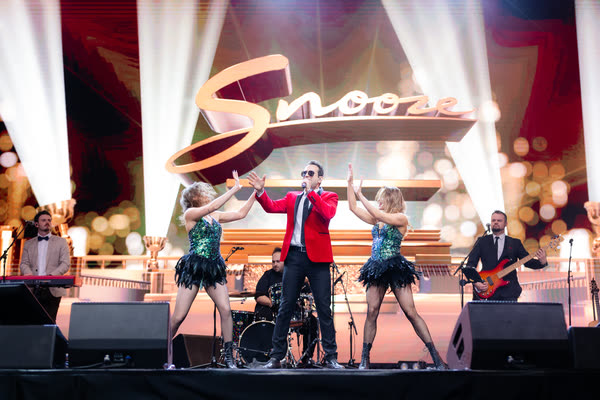 Sam in the red jacket singing with the band and two dancers in green sequins on a big gala stage