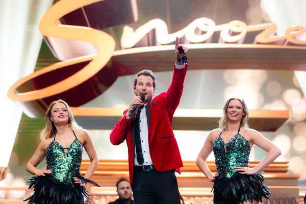 Sam in the red jacket singing and pointing to the crowd, a dancer in green sequins either side