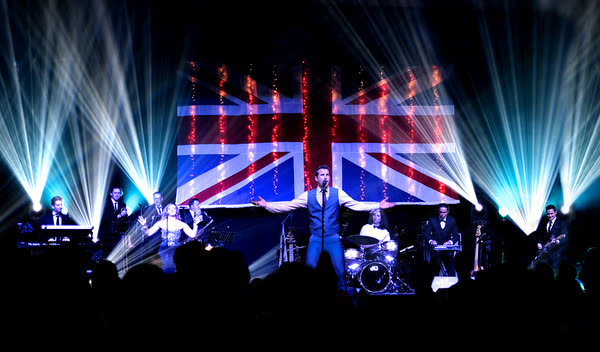 Sam with arms wide in front of a lit Union Jack backdrop, the full band and light beams around him