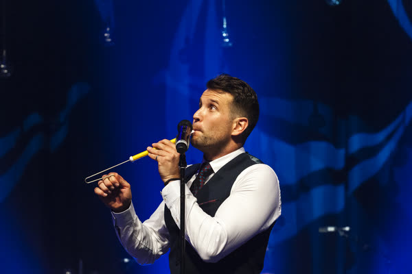Sam in a waistcoat playing a slide whistle into the microphone under blue stage light
