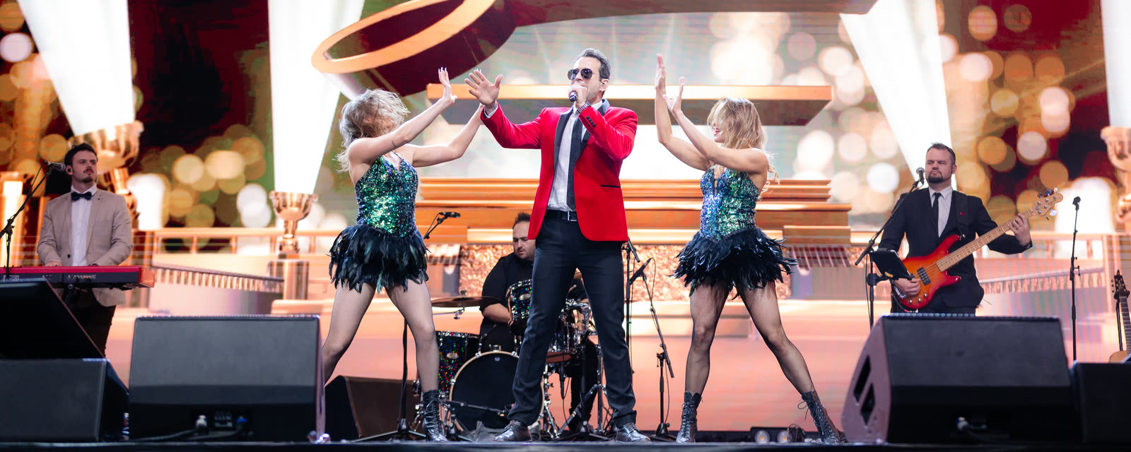 Sam in the red jacket singing on stage with the band and two dancers in green sequins